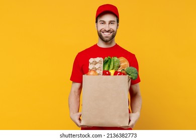 Smiling Delivery Guy Employee Man 20s Wear Red Cap T-shirt Uniform Workwear Work As Dealer Courier Hold Craft Brown Paper Bag With Grocery Food Vegetables Isolated On Plain Yellow Background Studio.