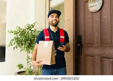 Smiling delivery man holding a smartphone and a paper bag delivering food to the front door of a house - Powered by Shutterstock