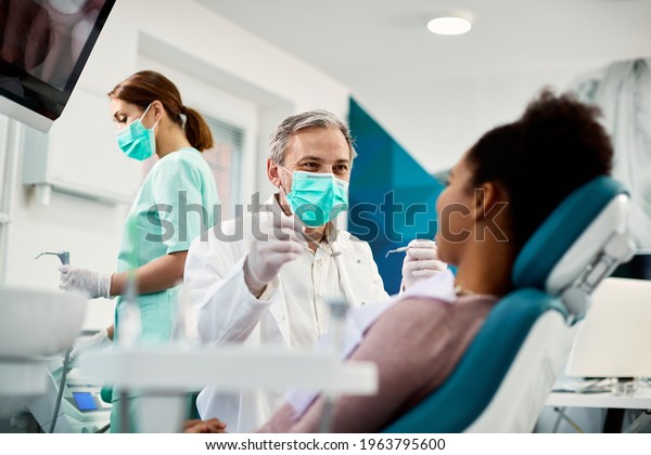 Smiling dentist communicating with African\
American woman while checking her teeth during dental procedure at\
dentist\'s office.