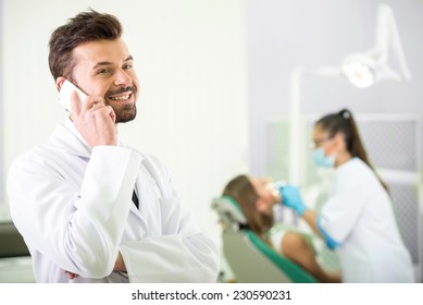 Smiling Dentist Is  Standing In His Office And Speaking By Phone. In The Background A Woman Dentist Treats A Patient.