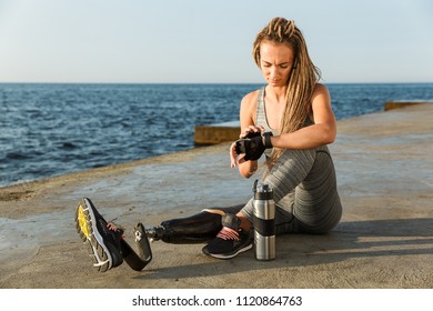 Smiling disabled athlete woman with prosthetic leg adjusting smartwatch while sitting at the beach - Powered by Shutterstock