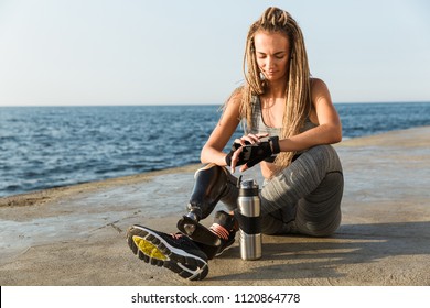 Smiling disabled athlete woman with prosthetic leg using smartwatch while sitting at the beach - Powered by Shutterstock