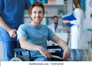 Smiling Disabled Young Man On Wheelchair At The Hospital, A Doctor Is Assisting Him