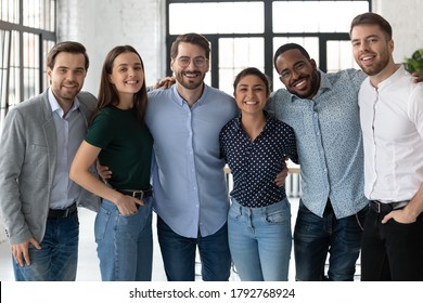 Smiling Diverse Business People, Successful Team, Staff Members Hugging, Standing In Modern Office, Looking At Camera, Happy Overjoyed Employees Colleagues Posing For Corporate Photo