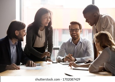 Smiling diverse businesspeople sit at office desk talk laugh discussing business ideas together, happy multiracial colleagues brainstorm joke negotiate at briefing in boardroom, cooperation concept