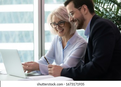 Smiling diverse colleagues sit at desk look at laptop screen watch funny webinar or video together, happy coworkers laugh working brainstorming discussing business ideas during briefing or meeting