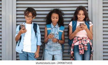 Smiling diverse multiethnic school kids children using smartphones and watching looking in mobile phone screen during break at the street outside standing over grey background - Powered by Shutterstock