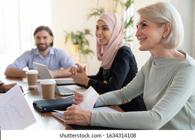 Smiling diverse multiracial office employees sit at desk cooperating at team meeting discussing business ideas together, happy multiethnic colleagues brainstorm talk at briefing, collaboration concept