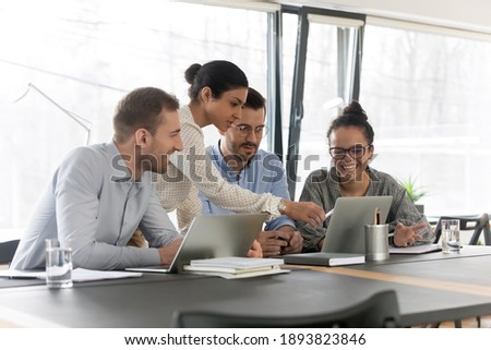 Smiling diverse young colleagues gather at desk in office look at laptop screen brainstorming at meeting together, happy multiracial employees cooperate using computer at briefing, teamwork concept