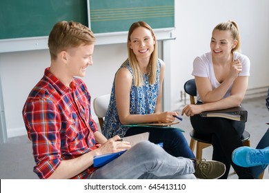 Smiling Diverse Young Students Having A Group Discussion In The Classroom In Front Of The Chalkboard Sitting In A Circle On Chairs