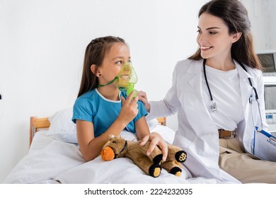 Smiling doctor with clipboard looking at happy child with oxygen mask in hospital - Powered by Shutterstock