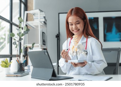 Smiling doctor explaining spinal anatomy using anatomical model of human skeleton during online consultation with patient - Powered by Shutterstock