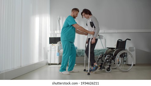 Smiling doctor helping young man patient with crutches. Portrait of medical worker in scrubs assisting injured man with broken leg during rehabilitation exercises in hospital - Shutterstock ID 1934626211