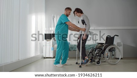 Smiling doctor helping young man patient with crutches. Portrait of medical worker in scrubs assisting injured man with broken leg during rehabilitation exercises in hospital
