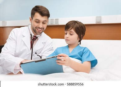 Smiling Doctor And Little Boy Patient Holding Clipboard On Hospital Bed