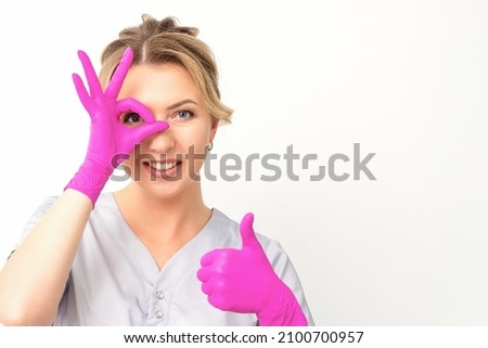 Smiling doctor oculist caucasian woman wearing pink rubber gloves in uniform showing ok sign covering the eye and thumb up gesture against a white background.