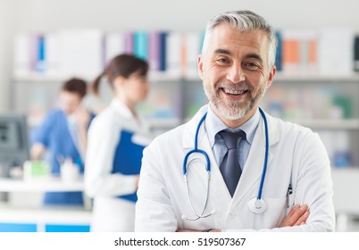 Smiling Doctor Posing With Arms Crossed In The Office, He Is Wearing A Stethoscope, Medical Staff On The Background