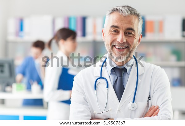 職場で腕を組んでポーズをとる微笑みを浮かべる医師 彼は聴診器を身に着け 背景に医療スタッフをつけている の写真素材 今すぐ編集