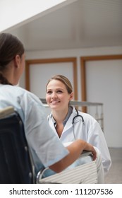 Smiling Doctor Squatting Next To A Patient On A Wheelchair In Hospital Corridor, Healthcare Workers In The Coronavirus Covid19 Pandemic