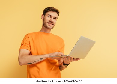 smiling dreamy man looking away while holding laptop on yellow background
