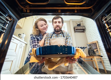 Smiling Eastern European Couple Cooking At Home. Man Taking Out A Pie From The Oven With Kitchen Interior And A Cat On Background, View From Inside The Oven.