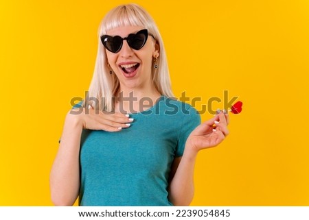 smiling eating a heart lollipop, blonde caucasian girl in studio on yellow background