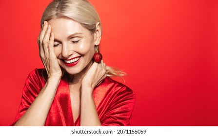 Smiling elder woman in beautiful Christmas gown with xmas tree bubble on ear, laughing glamourous, standing over red background in evening makeup - Powered by Shutterstock