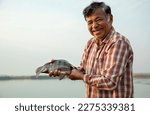 Smiling elderly Asian Fisherman holding a tilapia fish with lake on the background.
