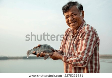 Smiling elderly Asian Fisherman holding a tilapia fish with lake on the background.