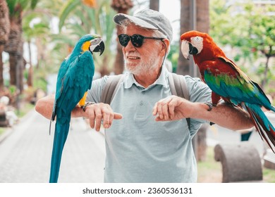 Smiling elderly bearded man in hat and sunglasses holding on his arms two domestic ara parrots in the park - Powered by Shutterstock