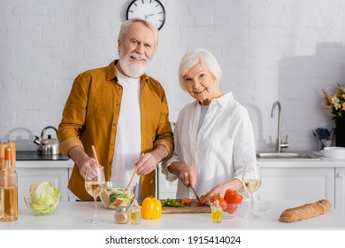 Smiling Elderly Couple Cooking Fresh Salad Near Wine And Baguette In Kitchen