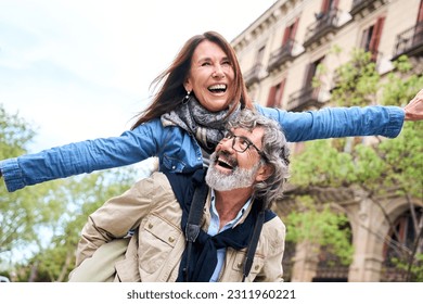 Smiling elderly couple having fun together on vacation. Older man piggybacking mature woman enjoying leisure time visiting European city. Concept of people in love relationships in retirement. - Powered by Shutterstock
