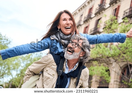 Smiling elderly couple having fun together on vacation. Older man piggybacking mature woman enjoying leisure time visiting European city. Concept of people in love relationships in retirement.