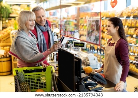 Smiling elderly couple paying with cell phone at supermarket, elderly man and woman embracing, standing by cashdesk, having conversation with friendly female cashier, using mobile phone