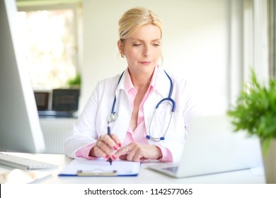 Smiling elderly female doctor sitting at doctor's room and using computer and laptop while writing diagnosis. 