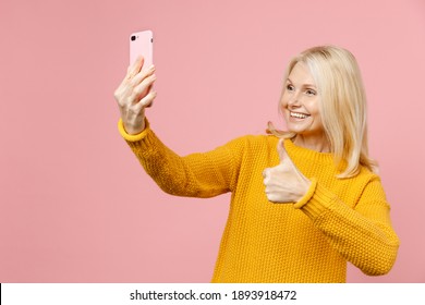 Smiling Elderly Gray-haired Blonde Woman Lady 40s 50s Years Old In Yellow Sweater Stand Doing Selfie Shot On Mobile Phone Showing Thumb Up Isolated On Pastel Pink Color Background Studio Portrait