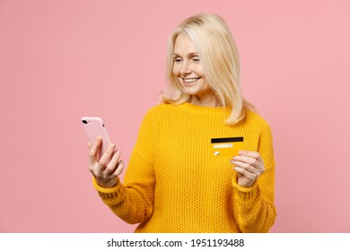 Smiling Elderly Gray-haired Blonde Woman Lady 40s 50s Years Old In Yellow Basic Sweater Standing Using Mobile Cell Phone Hold Credit Bank Card Isolated On Pastel Pink Color Background Studio Portrait