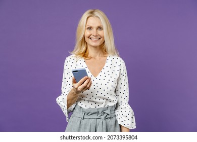 Smiling Elderly Gray-haired Blonde Woman Lady 40s 50s Years Old In White Dotted Blouse Using Mobile Cell Phone Typing Sms Message Looking Camera Isolated On Violet Color Background Studio Portrait