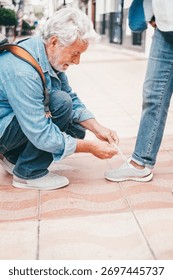 Smiling elderly man bends down to tie his wife's shoes. Two elderly people walking through the city enjoying free time