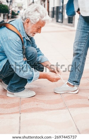 Smiling elderly man bends down to tie his wife's shoes. Two elderly people walking through the city enjoying free time