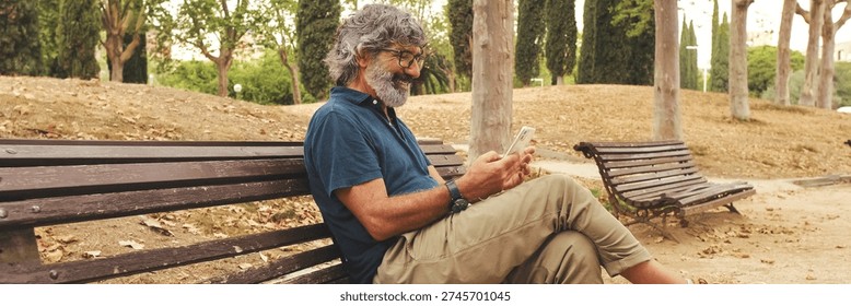 Smiling elderly man in glasses using phone while sitting on park bench. For banner. panoramic