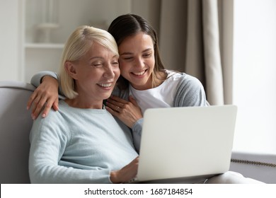 Smiling Elderly Mother And Adult Daughter Sit Relax On Sofa In Living Room Watch Video On Laptop Together, Happy Senior Mom And Grown-up Millennial Girl Child Rest At Home Using Modern Computer