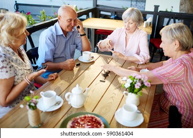 Smiling Elderly People Having Tea Party In Lovely Outdoor Cafe And Playing Dominoes Enthusiastically