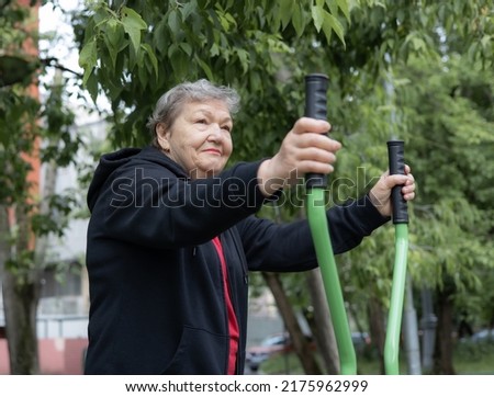 Smiling elderly woman is doing exercises on simulator outdoors in the yard.  Active life of pensioners. Adaptation of pensioners in the modern world. Prevention of Alzheimer's disease, sclerosis etc