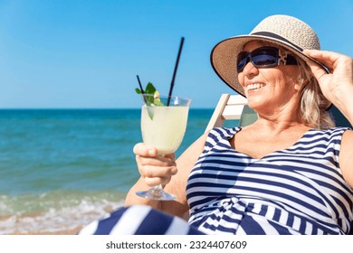 A smiling elderly woman in a dress and hat sits in a sun lounger on the seashore with a drink in her hand. Active lifestyle, travel, freedom and relax. Close-up. - Powered by Shutterstock