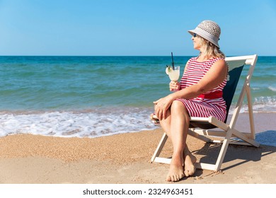 A smiling elderly woman in a dress and hat sits in a sun lounger on the seashore with a drink in her hand. Active lifestyle, travel and joy. - Powered by Shutterstock