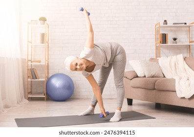 A smiling elderly woman is engaging in a fitness routine, holding a set of blue dumbbells while performing a side bend. She is standing on a yoga mat in a well-lit living room - Powered by Shutterstock