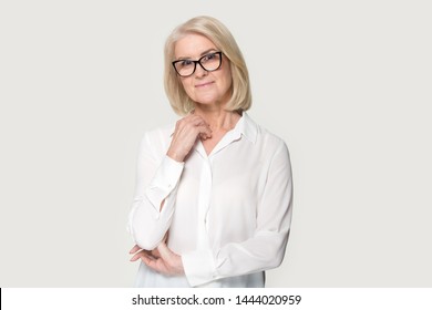 Smiling Elderly Woman In Glasses Feeling Shy Posing At Studio. Beautiful Old Lady Looking At Camera Feeling Uncertain, Unsure, Anxious. Studio Headshot Isolated On Grey Background. People Emotions