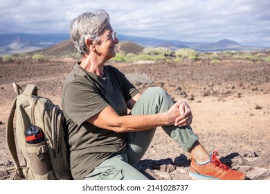 Smiling Elderly Woman On Outdoor Hike Sitting On A Stone Wall Looking Away. Active Mature Woman Sitting Near Her Backpack Enjoying Freedom And Sunny Day