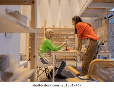 Smiling elderly woman shares her painted ceramic piece with a younger woman in a bright pottery studio. - Powered by Shutterstock